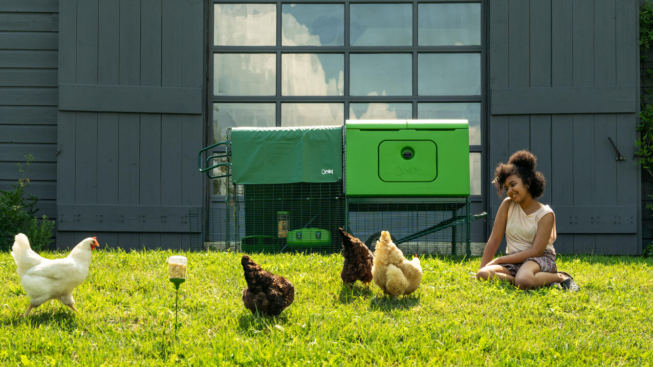 Mujer sentada en la hierba junto a su gallinero viendo a sus gallinas deambular