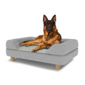 Perro sentado en una gran cama para perros Topology con almohadilla gris y patas redondas de madera