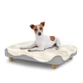 Perro sentado en una pequeña cama para perros Topology con topper de piel de oveja y patas redondas de madera