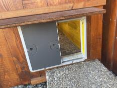 Un abridor de puerta de gallinero automático de color gris Omlet unido a un gallinero de madera.