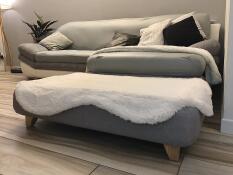 Una cama gris con un topper blanco de piel de oveja y pies cuadrados de madera